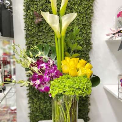 Tall arrangement of white calla lilies, green hydrangea, yellow roses, and purple orchids in a clear glass vase