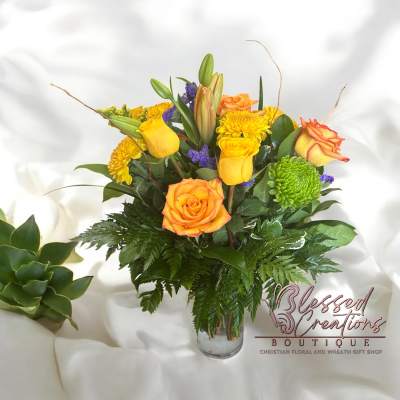 Bouquet of yellow and orange roses, mums, lilies, and purple accents in a clear glass vase