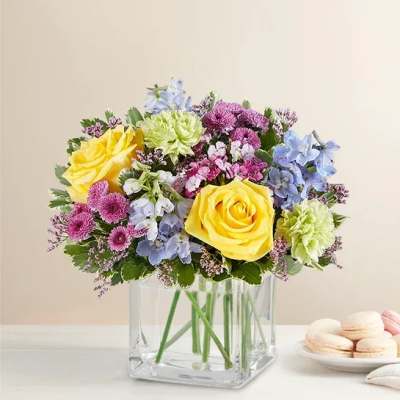 Bouquet of yellow roses, blue flowers, and pink blooms in a clear square vase