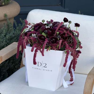Deep burgundy floral arrangement in a white gift bag with ribbon handles