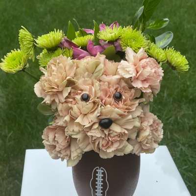 Dog-shaped peach carnation arrangement with green mums in a brown football vase on a white stand.