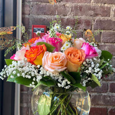 Round glass vase of pink, peach, and orange roses with white filler flowers and decorative butterflies.