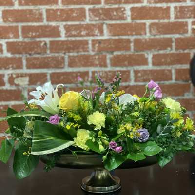 Low floral centerpiece with yellow roses, white lilies, and pink blooms in a pedestal bowl.