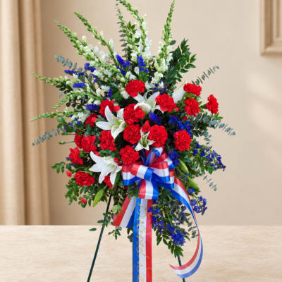 Standing floral spray with red carnations, white lilies, and blue accents on a ribboned easel