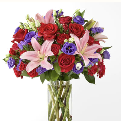 Bouquet of red roses, pink lilies, and purple blooms in a clear glass vase