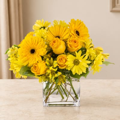Yellow gerberas and roses arranged in a clear square vase