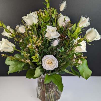 White roses arranged in a clear glass vase with greenery and small white accents