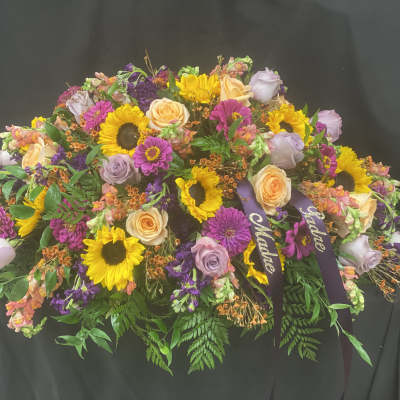 Casket spray with sunflowers, roses, and purple blooms on a dark drape