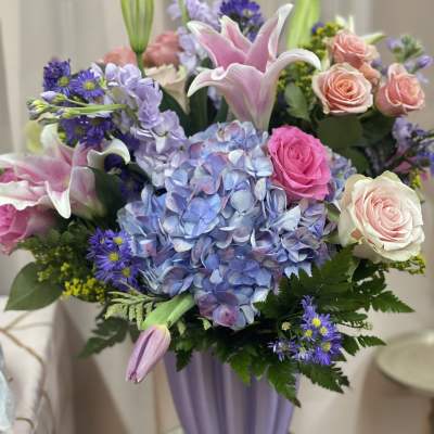 Pastel bouquet with hydrangea, roses, lilies, and purple filler flowers in a lavender vase