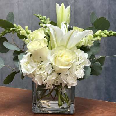 White lilies, roses, and hydrangeas arranged in a clear glass cube vase