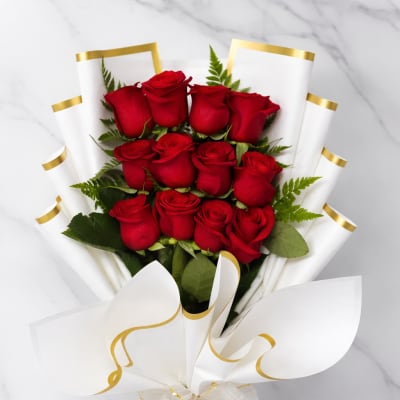 Hand-tied bouquet of red roses wrapped in white paper with gold trim and a decorative bow.