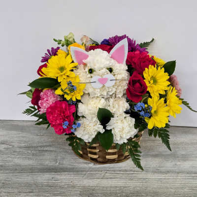 Basket arrangement with a white carnation cat face surrounded by yellow daisies, red roses, and bright mixed flowers