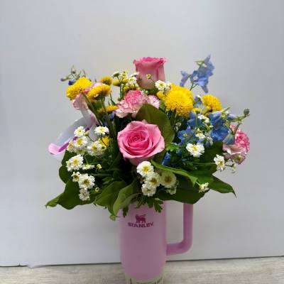 Mixed bouquet of pink roses, carnations, yellow mums, and blue blooms arranged in a pink tumbler.