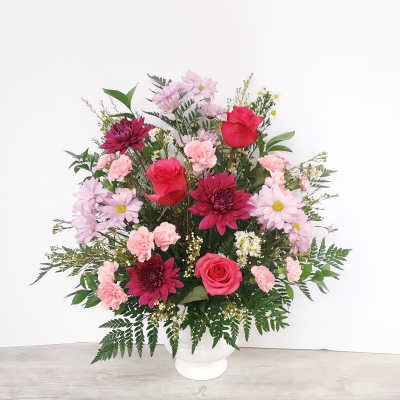 Tall arrangement of red roses, pink carnations, and mixed blooms in a white pedestal vase