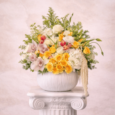 Yellow and white floral arrangement with roses, orchids, and tulips in a round white vase on a pedestal.