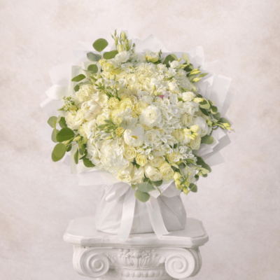 Round bouquet of white and pale yellow flowers wrapped in white fabric on a white pedestal.