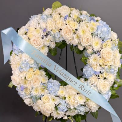 Standing wreath of white and pale blue flowers with a light blue message ribbon on an easel.