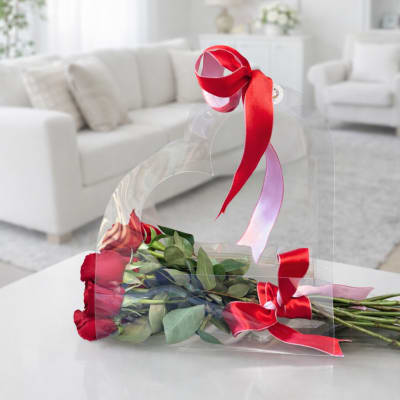 Bouquet of long-stem red roses in a clear heart-shaped carrier tied with red and pink ribbons on a white table
