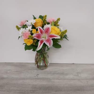 Mixed bouquet of pink lilies, yellow roses, white daisies, and carnations in a clear glass vase