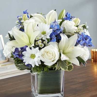 White lilies, roses, and blue flowers arranged in a square glass vase