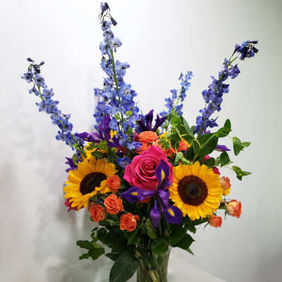 Mixed bouquet in a clear glass vase with sunflowers, roses, and blue delphinium