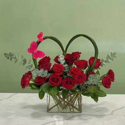 Red roses arranged in a glass vase with a heart-shaped frame and pink butterfly accents.