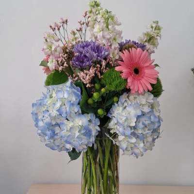 Mixed bouquet with blue hydrangeas, pink gerbera daisy, and purple blooms in a clear vase
