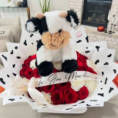 Round bouquet of red and white roses wrapped in cow-print paper with a plush cow toy on top.