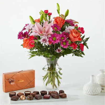 Mixed bouquet of pink lilies, roses, and daisies in a glass vase with chocolates