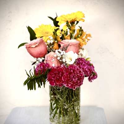 Mixed bouquet of pink roses, yellow daisies, and magenta carnations in a clear glass vase