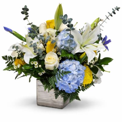 Mixed bouquet with white lilies, blue hydrangeas, and yellow roses in a square vase