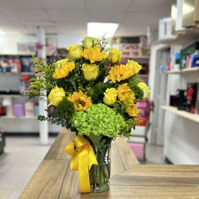 Yellow roses and sunflowers in a clear glass vase with a yellow ribbon