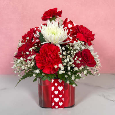 Red and white flower arrangement in a glass vase with heart ribbon