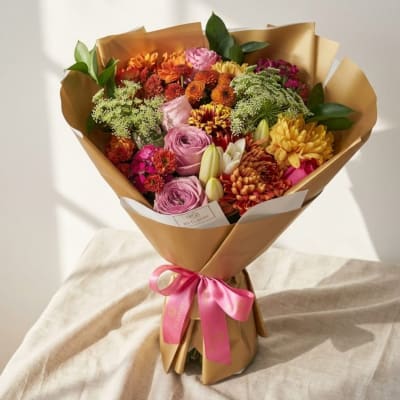 Mixed bouquet of pink, orange, and yellow flowers wrapped in brown paper with a pink ribbon