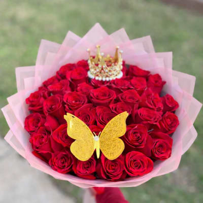 Handheld bouquet of red roses wrapped in pink paper with a gold butterfly and crown decoration.