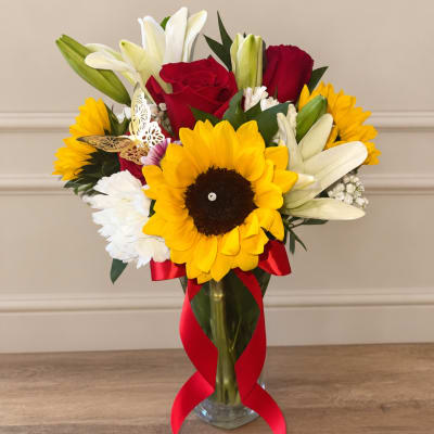 Bouquet of sunflowers, red roses, and white lilies in a glass vase with a red ribbon