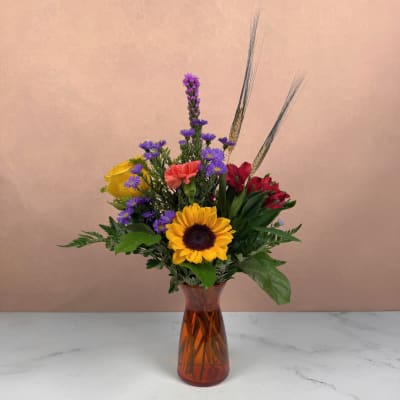 Mixed bouquet with a sunflower, roses, and purple daisies in an amber vase