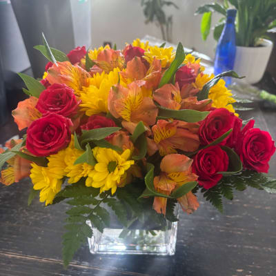 Bouquet of red roses, yellow daisies, and orange alstroemeria in a clear square vase