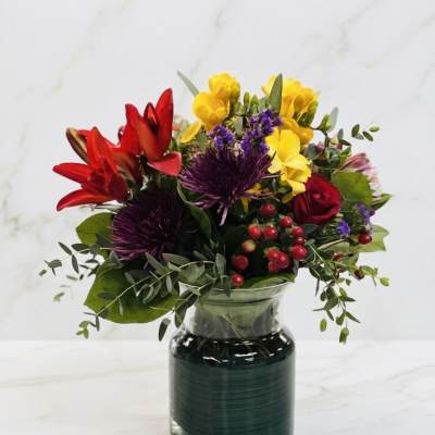 Bright mixed bouquet with red lilies, yellow blooms, purple mums and red roses in a clear glass vase