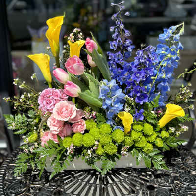 Colorful mixed flower arrangement with tulips, calla lilies, and blue delphinium in a white container