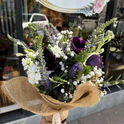 Handheld bouquet of purple, white, and green flowers wrapped in burlap