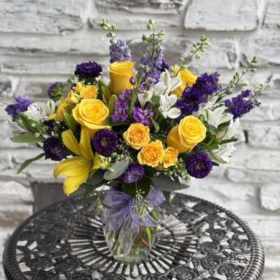 Bouquet of yellow roses, purple flowers, and white lilies in a glass vase