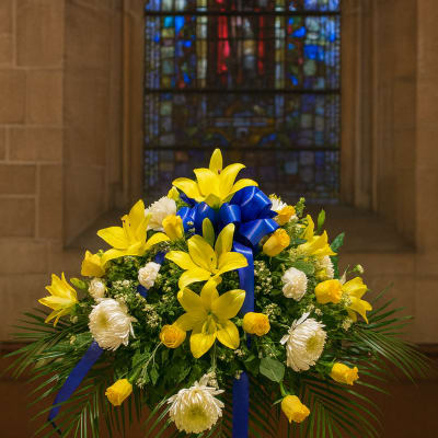 Yellow lilies and white flowers arranged with a blue ribbon on a pedestal.
