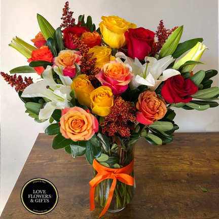 Tall vase arrangement of red, yellow, and orange roses with white lilies and an orange ribbon bow.