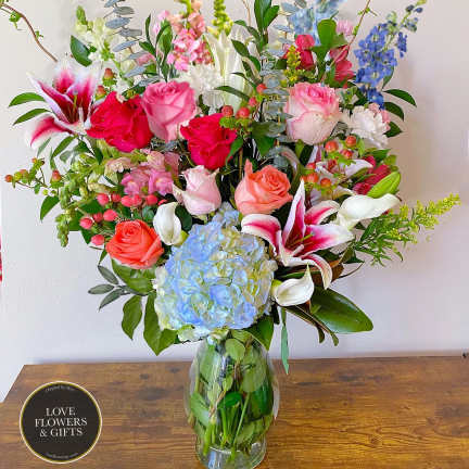 Tall mixed arrangement of pink roses, lilies, blue hydrangea and assorted blooms in a clear glass vase