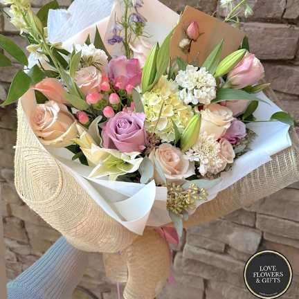 Hand-tied bouquet of pastel roses, lilies, and mixed blooms wrapped in white and burlap-style paper
