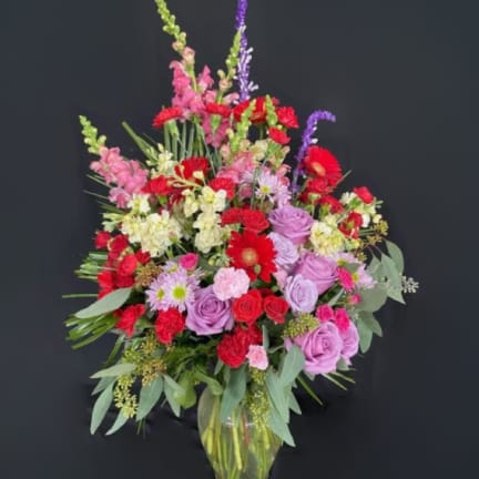 Tall mixed arrangement of red, pink, yellow, and lavender flowers in a clear glass vase