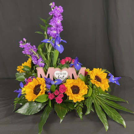 Colorful floral arrangement with sunflowers, purple blooms, and pink roses around a "Mom" sign