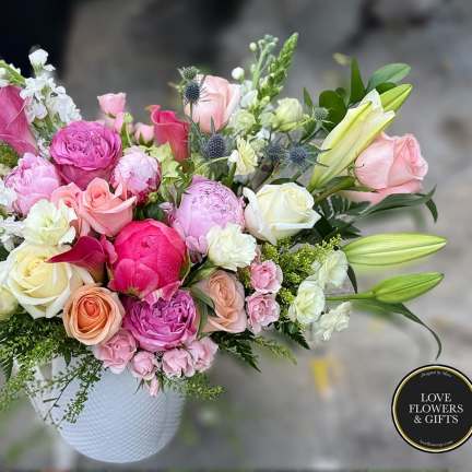 Mixed arrangement of pink peonies, roses, and lilies in a white ceramic vase