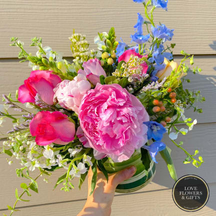 Bright bouquet of pink peonies, roses, and mixed blooms in a clear glass vase held in one hand